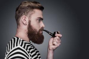 Confident sailor. Side view of confident young bearded man in striped clothing smoking a pipe and looking away while standing against grey background photo