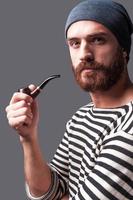 Me and my pipe. Side view of confident young bearded man in striped clothing holding a smoking pipe and looking at camera while standing against grey background photo