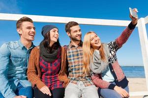 Capturing a happy moment. Low angle view of four happy young people bonding to each other and making selfie while sitting at the riverbank photo