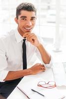 agradable trabajador guapo joven afroamericano en camisa y corbata tomados de la mano en la barbilla y sonriendo mientras está sentado en su lugar de trabajo foto