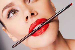 Cosmetics artistry. Close-up of a beautiful woman with red lips holding make-up brush in her mouth and looking away while standing against grey background photo