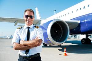 Confident pilot. Confident male pilot in uniform keeping arms crossed and smiling with airplane in the background photo