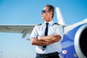 apasionado por el cielo. piloto masculino confiado en uniforme manteniendo los brazos cruzados y mirando hacia otro lado con un avión en el fondo foto