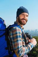 On the top. Handsome young man carrying backpack and looking at camera through the shoulders with smile while standing in the nature and holding binoculars photo