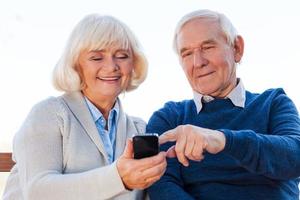 Technologies become easier. Cheerful senior couple looking at the mobile phone and smiling while sitting on the park bench together photo