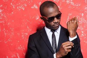 Everything should be perfect. Serious young African man in formalwear and sunglasses adjusting his sleeve while standing against red background photo