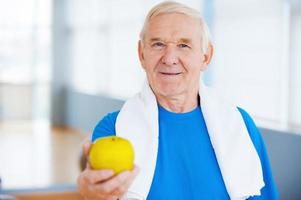 Join healthy lifestyle Happy senior man with towel on shoulders stretching out green apple while standing in health club photo