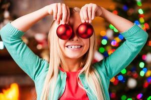 Christmas fun. Cute little girl holding Christmas balls in front of her eyes and smiling with Christmas Tree and fireplace in the background photo