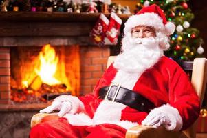 ho ho ho tradicional santa claus sentado en su silla y sonriendo con chimenea y árbol de navidad en el fondo foto