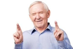 Waiting for special moment. Portrait of senior man in shirt keeping fingers crossed and eyes closed while standing against white background photo