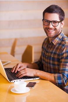 Having opportunity to work everywhere. Handsome young man working on laptop and smiling while sitting in cafe photo