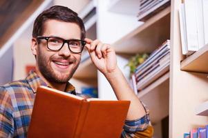 I love studying. Handsome young man holding book and smiling while standing in front of the bookshelf photo