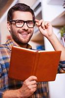 Handsome bookworm. Handsome young man holding book and smiling while standing in front of the bookshelf photo
