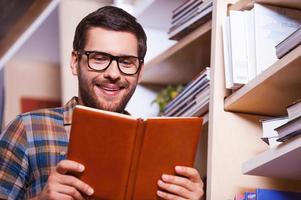 I love this book. Handsome young man reading book and smiling while standing in front of the bookshelf photo