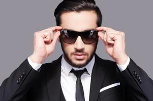 I put the man in manly. Portrait of handsome young man in formalwear adjusting his sunglasses while standing against grey background photo