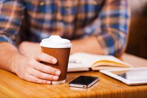 tomando tiempo para el descanso para tomar café. primer plano de un hombre que sostiene una taza de café mientras está sentado en el escritorio con un bloc de notas y una tableta digital sobre él foto