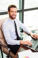 Confident businessman. Top view of handsome young man in shirt and tie looking at camera and smiling while sitting at his working place photo