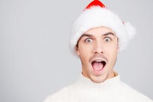Unbelievable Surprised young man in Santa hat keeping mouth open and looking at camera while standing against grey background photo