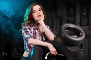 Beautiful auto mechanic. Beautiful young woman leaning at the car tire and looking at camera while sitting in auto repair shop photo