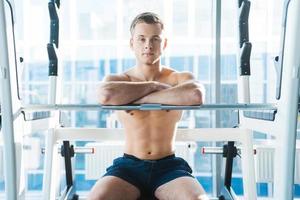 Resting after workout session. Confident young muscular man keeping arms crossed and looking at camera while sitting at the bench press photo
