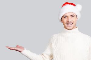 Copy space in my hand. Handsome young man in Santa hat holding copy space and smiling while standing against grey background photo