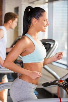 Treadmill workout. Side view of attractive young woman running on a treadmill and smiling with man exercising in the background photo