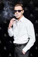 fumando guapo. un joven apuesto con camisa blanca fumando cigarrillos y mirando a la cámara mientras se enfrenta a un fondo negro foto