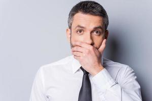 I am shocked Surprised mature man in shirt and tie covering mouth with hand and looking at camera while standing against grey background photo