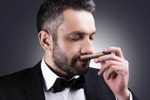 Great cigar Portrait of handsome mature man in formalwear smelling cigar and keeping eyes closed while standing against grey background photo