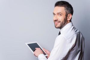 Man with digital tablet. Rear view of happy mature man in shirt and tie working on digital tablet and looking over shoulder while standing against grey background photo
