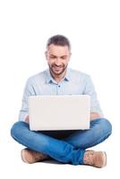 Man surfing the net. Handsome young man in casual wear sitting on the floor and working on laptop while being isolated on white background photo