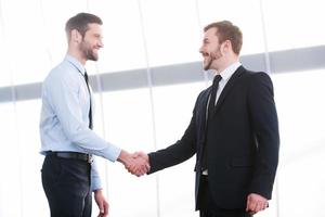 sellando un trato. dos alegres hombres de negocios dándose la mano y sonriendo mientras están parados adentro foto