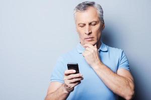 Choosing the right words. Serious mature man in T-shirt holding mobile phone and looking at it while standing against grey background photo
