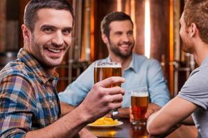 Spending time with friends. Handsome young man toasting with beer and smiling while sitting with his friends in beer pub photo