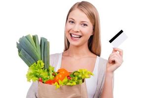 Verduras frescas que compro todos los días. hermosa mujer joven que lleva una bolsa de compras llena de comestibles y muestra su tarjeta de crédito mientras está de pie contra el fondo blanco foto