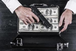 Illegal business. Top view of man holding gun and opening a briefcase full of paper currency photo