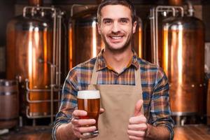 cervecero feliz. feliz joven cervecero en delantal sosteniendo un vaso con cerveza y mirándolo con una sonrisa mientras está parado frente a contenedores de metal foto