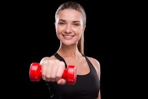 Join healthy lifestyle Sporty young woman holding dumbbell and smiling while standing against black background photo