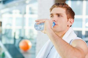 refrescante después del ejercicio. joven cansado que lleva una toalla en los hombros y bebe agua mientras está de pie en el gimnasio foto