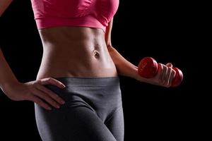 Perfect body. Close-up of beautiful young woman with fit body holding dumbbell while standing against black background photo