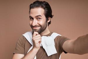 Handsome young man making selfie while standing against brown background photo