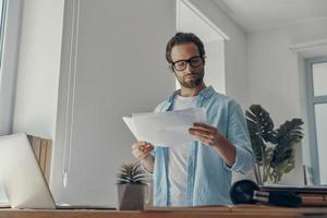 Concentrated young man doing some paperwork while standing near his working place in office photo