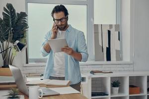 Confident man talking on phone and looking at the digital tablet while standing in office photo