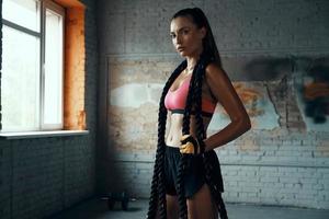 Beautiful young woman carrying battle rope and looking at camera while standing in gym photo