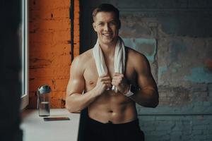 Handsome muscular man carrying towel on shoulders and smiling while standing in gym photo
