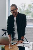 Cheerful young man using laptop while standing near his working place in office photo