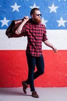 viajando con estilo. longitud completa de un apuesto joven barbudo con gafas de sol dando un paso y sosteniendo una maleta en su hombro contra la bandera americana foto