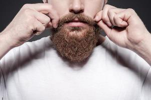haciendo su propio estilo. primer plano de un joven barbudo ajustando sus bigotes mientras se enfrenta a un fondo gris foto