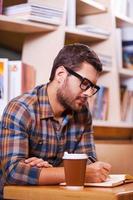 Spending time in library. Confident young man writing something in note pad while sitting at the desk in library photo