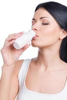 Small bottle but  so much usefulness. Beautiful young smiling woman drinking from white little bottle while standing against white background photo